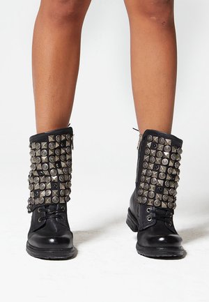 Botines negros hasta el tobillo con tachuelas plateadas decorativas y detalles en forma de pirámide, cierre con cordones en la parte delantera y cremallera en el lateral.