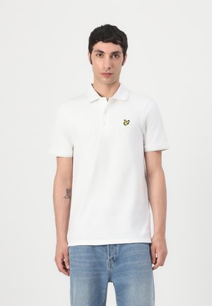 Vit bomullspolo med korta ärmar, tvåknäppning och en gul broderad logotyp på vänster bröst. Klassisk figursydd design.