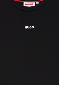 Svart t-skjorte laget av bomull, med rund hals og liten hvit 'HUGO'-logo i midten. Den indre kragen har en rød detalj.