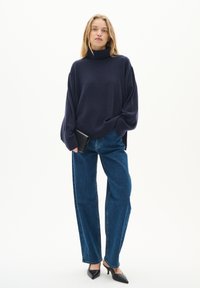 Marinefarbener Rollkragenpullover mit übergroßen Ärmeln, kombiniert mit hoch taillierten blauen Jeans und schwarzen, spitzen Pumps, während sie eine schwarze Clutch hält.