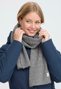 Kvinde i en navy-blå jakke smiler, mens hun justerer et blødt gråt strikket tørklæde, der er viklet om hendes hals, mod en ensfarvet lys baggrund.