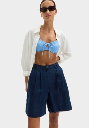 Kvinde iført sorte solbriller, hvid åben skjorte, lyseblå cropped top og marineblå højtaljede plisserede shorts, stående med den ene hånd på hoften.