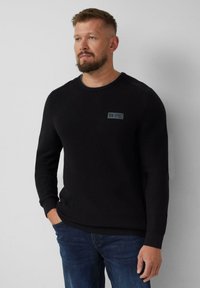 Homme avec une barbe portant un pull noir texturé et un jean bleu, se tenant avec une main dans la poche contre un fond clair.