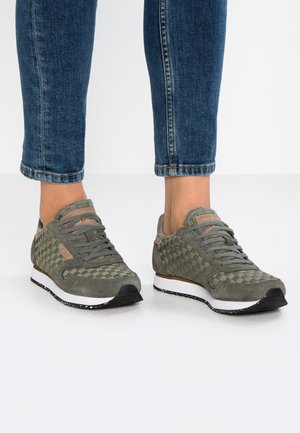 Person iført grønne vævede sneakers med hvide såler og blå jeans, der står på en hvid gulv.