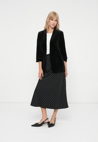 Fekete bársony blazer egy fehér felső fölött, hosszú fekete pöttyös szoknyával párosítva. Fekete slingback cipők hegyes orral.