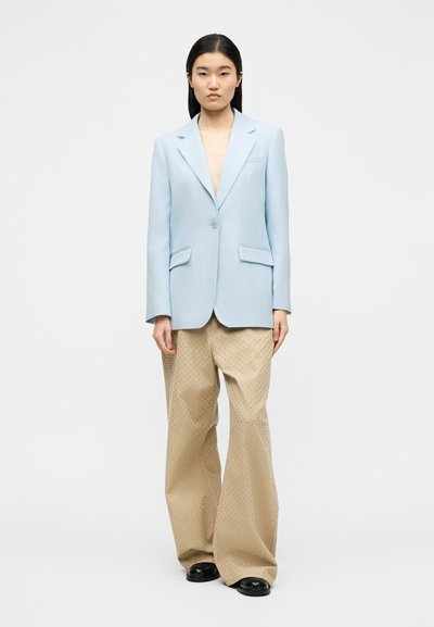 Γυναίκα που φοράει ανοιχτό μπλε σακάκι κουμπωμένο μία φορά, μπεζ παντελόνι με φαρδιά μπότα και μικρές τρύπες, και μαύρα παπούτσια, στέκεται μπροστά σε λευκό φόντο.