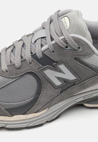 La chaussure de sport grise présente un mélange de matériaux en daim et en mesh, un dessus texturé et un logo argenté proéminent sur le côté.