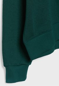 Gros plan sur le poignet et l’ourlet d’un sweat vert foncé, montrant la texture côtelée et les détails des coutures sur un fond uni.