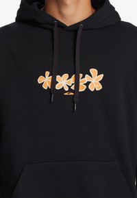 Sudadera con capucha negra con gráfico floral naranja y cordones, recortada a la altura del cuello y el pecho, mostrando parte de un bolsillo tipo canguro.