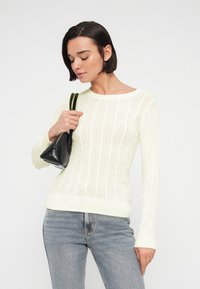 Vrouw met kort donker haar, gekleed in een crèmekleurige kabelgebreide trui en lichtblauwe spijkerbroek, met een kleine zwarte handtas op haar schouder.