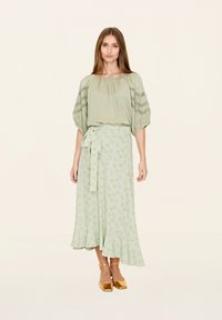 Helles grünes Blusenshirt mit Spitzenverzierungen, kombiniert mit einem langen, gemusterten Rock mit floralen Motiven und goldenen Blockabsatzschuhen.