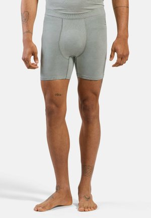 Male wearing gray fitted boxer briefs, standing barefoot, showing various tattoos on legs and arms against a plain white background.