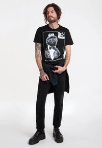 Camiseta negra con diseño del Monstruo de las Galletas en blanco, combinada con pantalones negros y botas negras gruesas. Tatuajes visibles en los brazos.