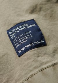 Fondo de tela verde con una etiqueta azul cosida sobre ella. La etiqueta muestra el nombre de la marca "Superdry", instrucciones de cuidado y una advertencia de no retirarla.