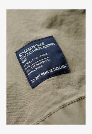 Groene stoffen achtergrond met een blauwe label erop gestikt. Het label toont de merknaam "Superdry," onderhoudsinstructies en een waarschuwing om het niet te verwijderen.