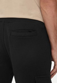Pantaloni della tuta neri con finitura testurizzata, dotati di una tasca posteriore e di un elastico in vita. Il tessuto appare morbido e progettato per il comfort.