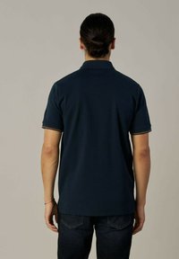 Tumesinine polo särg lühikeste varrukatega, ribatatud krae ja kontrastsete triipudega varrukate äärestel, valmistatud siledast ja hingavast kangast.