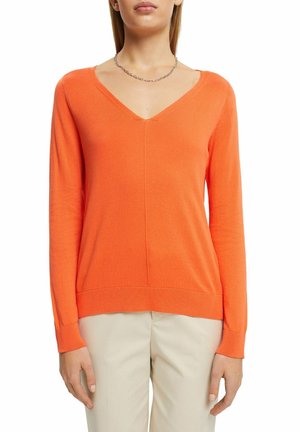 Femme portant un pull orange vif à manches longues et col en V, avec un pantalon beige et un collier en chaîne argentée, sur fond blanc.