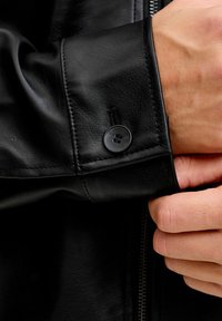 Schwarzer Lederjackenärmel mit glatter Textur, ausgestattet mit einem Knopfverschluss und sichtbaren Nahtdetails am Ärmelabschluss. Vorderseite mit Reißverschluss.