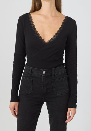 Femme portant un haut cache-cœur noir à manches longues avec bordure en dentelle et un jean noir taille haute avec poches plaquées devant sur fond uni.