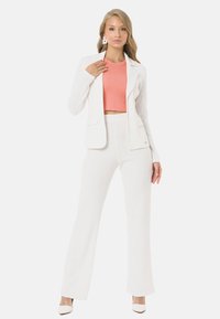 Weißes gewebtes Anzug-Set mit einer taillierten Blazerjacke und ausgestellten Hosen, drunter einem korallenfarbenen Cropped-Top. Schlichte Ohrringe runden den Look ab.