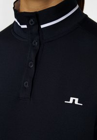 Schwarzes Polohemd mit hohem Kragen, ausgestattet mit einem weißen Streifen, Knopfleiste und einem kleinen weißen Logo auf der Brust. Glatte Stofftextur.