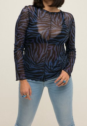 Mujer con una blusa azul y negra de rayas de cebra, transparente y de manga larga, con vaqueros azul claro y varios anillos en los dedos.