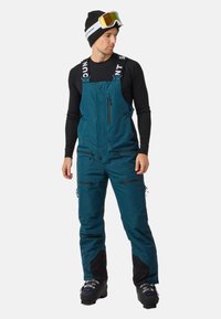 Groene skibroek met verstelbare banden, ritsen op de borst en benen, versterkte zwarte onderbenen, gedragen over een zwart lang T-shirt.