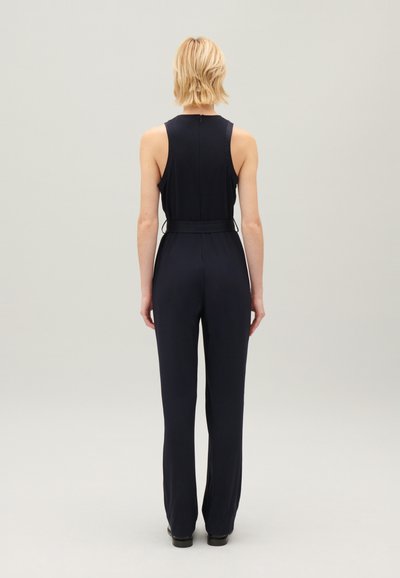 Combinaison navy avec une taille cintrée, un design sans manches et un dos ouvert. Tissu lisse, jambes droites et accents matériels minimalistes.