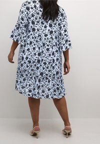 Himmelblaues Kleid mit schwarzem Blumenmuster, ausgestellt geschnittenen Ärmeln, gestuftem Rüschensaum und fließendem Design. Getragen mit beigen, hochhackigen Sandalen.