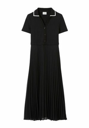 Femme portant une robe noire à manches courtes avec un liseré blanc au col, tenant un petit sac à main marron et portant des sandales dorées à brides.