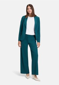 Teal blazer en wijde broek gecombineerd met een witte top. Gladde stof, op maat gemaakt ontwerp en scherpe revers. Geaccentueerd met spitse beige hakken.