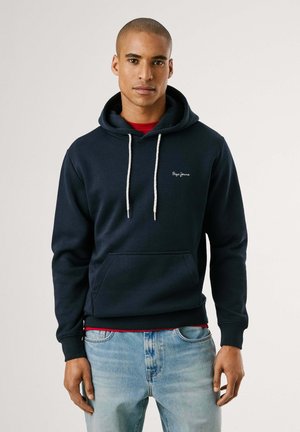 Navyblauwe hoodie van zachte stof, met een kangoeroezak, een capuchon met trekkoord en een geborduurd logo. Draagt over een rood shirt.