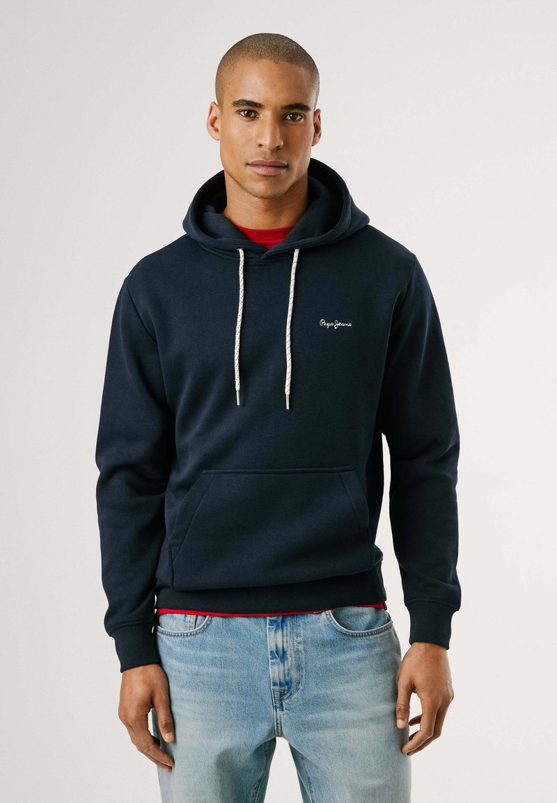 Sudadera azul marino de tela suave, con un bolsillo canguro, capucha con cordón y logo bordado. Usada sobre una camiseta roja.