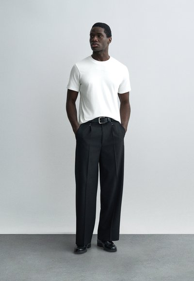 Homme debout, mains dans les poches, portant un t-shirt blanc, un pantalon large noir, une ceinture noire et des chaussures habillées noires, sur un fond uni.