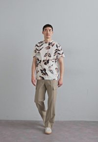 Hvid skjorte med abstrakte brune og sorte blomster mønstre, parret med beige cargo bukser. Modellen står på et gråt gulv foran en grå væg.