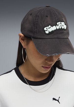 Donna che indossa un cappellino da baseball nero consumato con la scritta "Fun A On", grandi orecchini a cerchio, collana d'argento e una maglietta bianca Puma con rifiniture nere.
