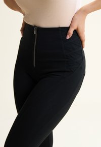 Hochtaillierte schwarze Leggings aus dehnbarem Stoff. Verfügt über einen Reißverschluss vorne und ein strukturiertes Paneel mit diagonalen Linien an den Seiten.