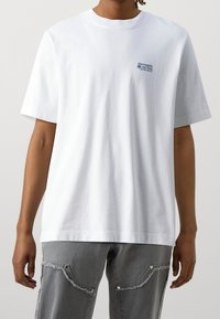 Person iført en enkel hvid t-shirt med korte ærmer, der har lille blå tekst på brystet, parret med grå jeans med flossede kanter.