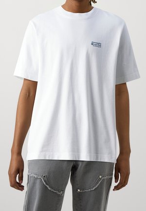 Personne portant un t-shirt blanc à manches courtes uni avec un petit texte bleu sur la poitrine, associé à un jean gris avec des bords effilochés.