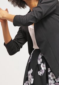 Černý přizpůsobený blazer s dlouhými rukávy, vrstvený přes světlý top, spárovaný s květinovou midi suknią s fialovými a bílými vzory.