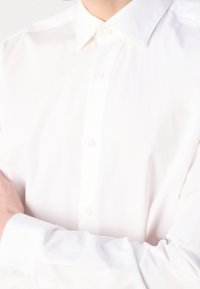 Camicia bianca con bottoni e colletto classico. Tessuto liscio, con bottoni perlati. Maniche lunghe con polsini visibili.