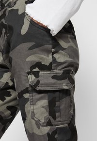 Tarnfarbenes Cargo-Hose in verschiedenen Grautönen und Schwarz. Verfügt über eine seitliche Tasche mit Klappe und ein distressed Bündchen an den Ärmeln.