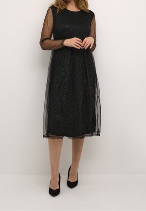 Vrouw draagt een knielange zwarte jurk met doorschijnende lange mouwen en kleine witte stippen, gecombineerd met zwarte pumps met spitsen neus.