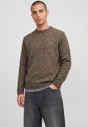 Homme aux cheveux courts portant un pull en maille marron tacheté, une chemise bleue en dessous et un jean gris foncé, debout avec une main dans la poche.