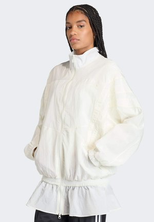 Mujer con el cabello trenzado, vistiendo una chaqueta blanca suelta con cremallera, puños elásticos y dobladillo con volantes, de pie con las manos en los bolsillos.