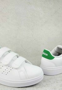 Baskets blanches avec deux bandes Velcro, design perforé sur les côtés et une accentuation verte au niveau du talon avec le logo "adidas". Semelle en caoutchouc.