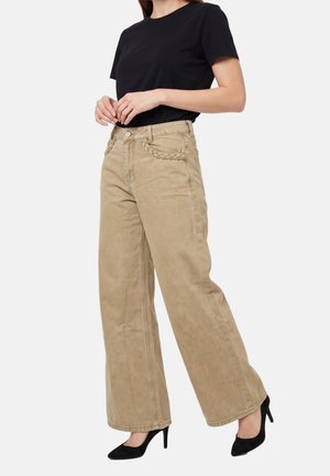 Donna che indossa pantaloni beige a gamba larga con dettagli intrecciati sulle tasche, top nero a maniche corte e scarpe nere con tacco alto, in piedi con le mani giunte.