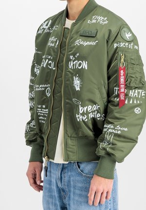 Personne portant une veste bomber verte avec un texte et des symboles style graffiti blancs promouvant la paix, l'amour et le respect à l'avant et sur les manches.