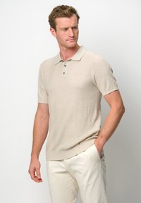 Beige kortärmad pikétröja med ett texturerat mönster, två knappar och en klassisk krage. Bärs med ljusa byxor.